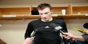 Cale Makar in a black adidas workout shirt looking down while surrounded by microphones