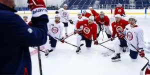 A group of Capitals rookies kneel on the ice in front of a coach.